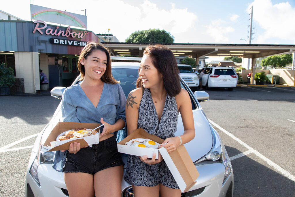 Two friends seated on the hood of a Toyota Prius enjoying plate lunches in front of Rainbow Drive In discussing how to save money as a college student on Oahu.